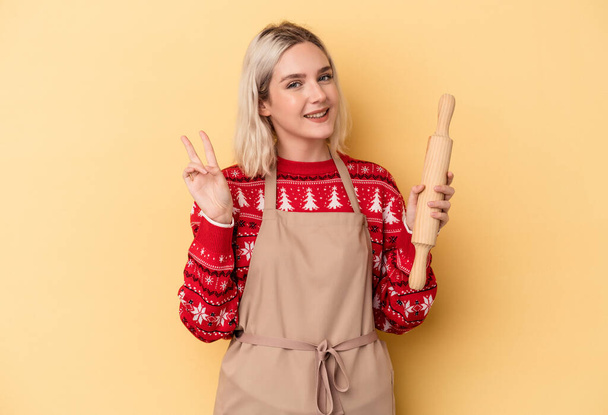 Νεαρή λευκή φουρνάρισσα που φτιάχνει μπισκότα για τα Χριστούγεννα απομονωμένη σε κίτρινο φόντο χαρούμενη και ανέμελη δείχνοντας ένα σύμβολο ειρήνης με δάχτυλα. - Φωτογραφία, εικόνα