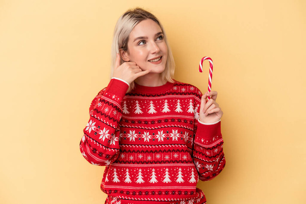 Young caucasian woman holding a christmas stick isolated on yellow background showing a mobile phone call gesture with fingers. - Photo, Image