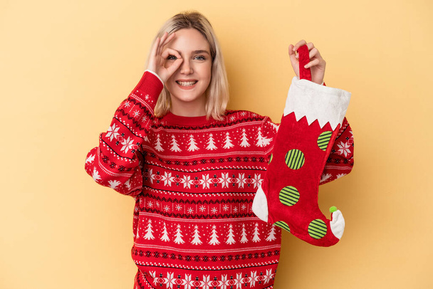 Young caucasian woman holding a elf sock isolated on yellow background excited keeping ok gesture on eye. - Photo, Image