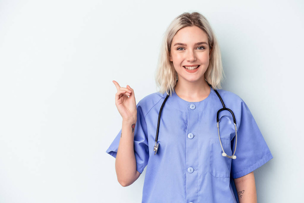 Jeune infirmière femme isolée sur fond bleu souriant et pointant du doigt, montrant quelque chose à vide. - Photo, image