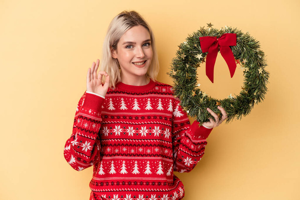 Joven mujer caucásica sosteniendo una corona de Navidad aislada sobre fondo amarillo alegre y confiado mostrando gesto ok. - Foto, Imagen
