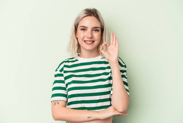 Молодая белая женщина, изолированная на зеленом фоне, подмигивает глазом и держит нормальный жест рукой.. - Фото, изображение