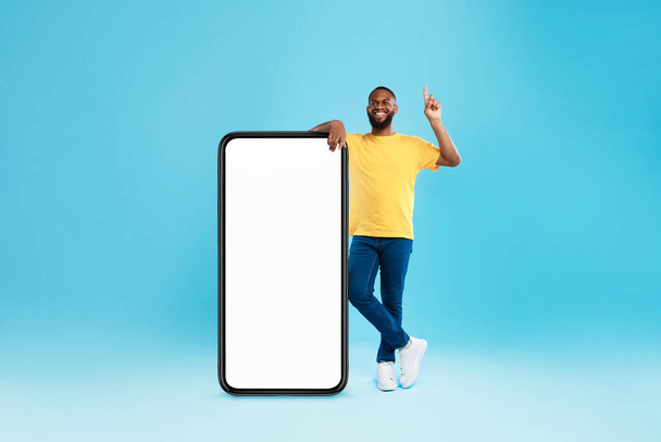 Handsome black guy offering mockup for new mobile app on giant smartphone screen, pointing upwards on blue background - Photo, Image