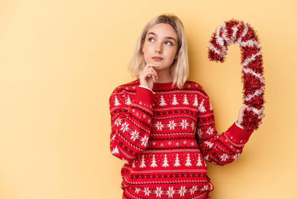 Mladá běloška drží velké vánoční hůl izolované na žlutém pozadí dívá bokem s pochybným a skeptickým výrazem. - Fotografie, Obrázek