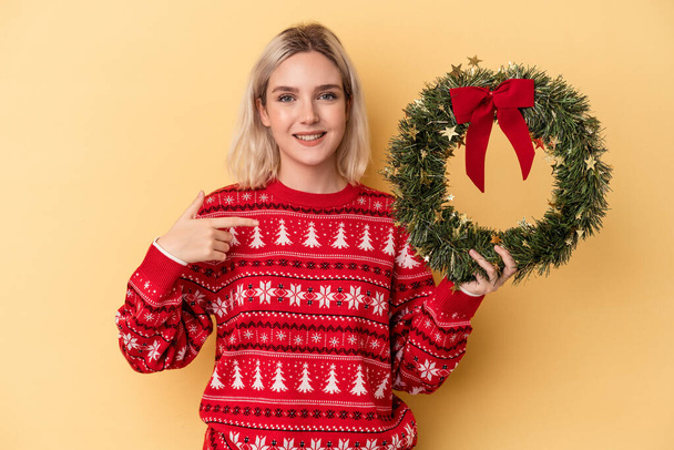 Jonge blanke vrouw met een kerstkrans geïsoleerd op gele achtergrond persoon wijzend met de hand naar een shirt kopieerruimte, trots en vol vertrouwen - Foto, afbeelding
