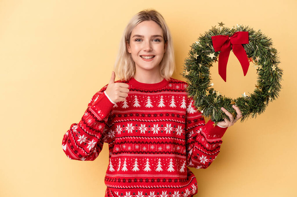 Young caucasian woman holding a Christmas wreath isolated on yellow background smiling and raising thumb up - Photo, Image