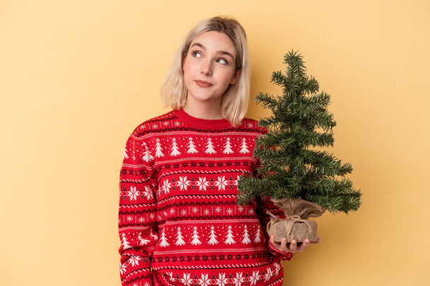 Joven mujer caucásica sosteniendo un pequeño árbol de navidad aislado sobre fondo amarillo soñando con lograr metas y propósitos - Foto, imagen