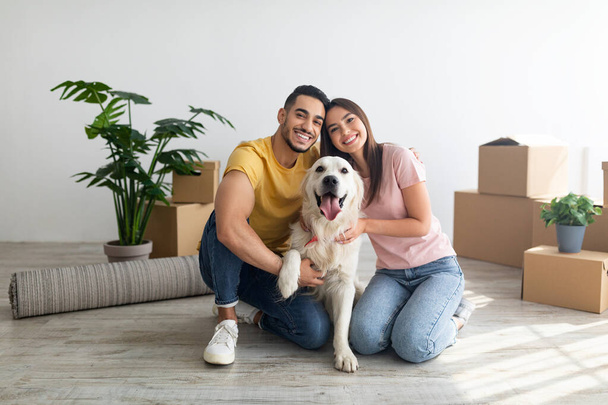 Cheery young international couple with cute golden retriever dog sitting on floor of new home on relocation day - Fotó, kép