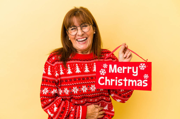 Mujer caucásica de mediana edad sosteniendo alegre cartel de Navidad aislado sobre fondo amarillo riendo y divirtiéndose.  - Foto, Imagen