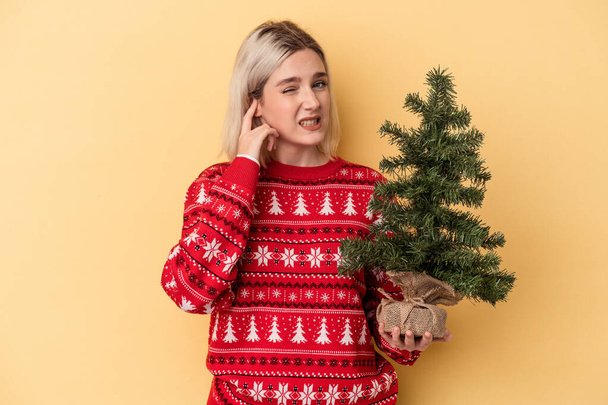 Молодая белая женщина держит маленькую рождественскую елку на желтом фоне, закрывая уши руками. - Фото, изображение