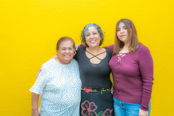 Drei Generationen lateinamerikanischer Frauen lächeln, Großmutter, Tochter und Enkelin tragen lässige Kleidung und blicken in die Kamera mit gelbem Hintergrund - Foto, Bild