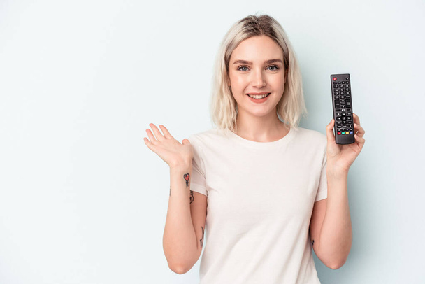 Mulher caucasiana jovem segurando um controlador de tv isolado no fundo azul mostrando um espaço de cópia em uma palma e segurando outra mão na cintura. - Foto, Imagem