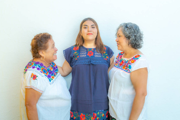 Moeder en dochter kijken naar elkaar en kleindochter in het midden van hen, drie generaties Mexicaanse vrouwen glimlachend met bloemenprint blouses op een witte achtergrond - Foto, afbeelding