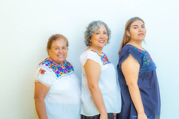 Three generations of smiling Mexican women with floral print blouses in a row looking at the camera on a white background - Photo, Image