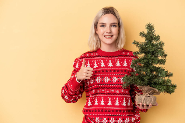 Mladá běloška žena drží malý vánoční stromek izolované na žlutém pozadí s úsměvem a zvednutím palce nahoru - Fotografie, Obrázek
