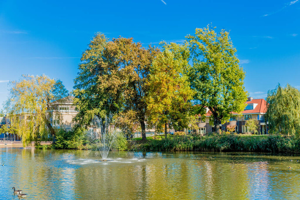 Pond with a fountain running with slight reflection in the water, lush trees with yellow green foliage and houses in the background, sunny day in early autumn in Voerendaal, South Limburg, Netherlands - Zdjęcie, obraz