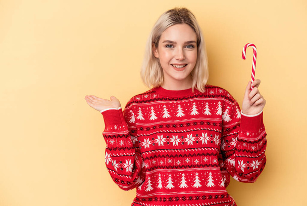 Young caucasian woman holding a christmas stick isolated on yellow background showing a copy space on a palm and holding another hand on waist. - Photo, Image