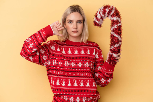 黄色の背景に孤立した大きなクリスマススティックを持つ若い白人女性は嫌いなジェスチャー、親指を示しています。意見の相違. - 写真・画像