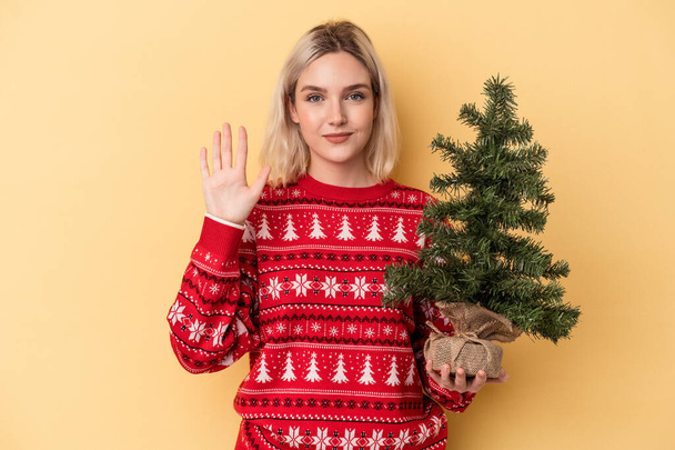 Joven mujer caucásica sosteniendo un pequeño árbol de navidad aislado sobre fondo amarillo sonriente alegre mostrando el número cinco con los dedos. - Foto, imagen