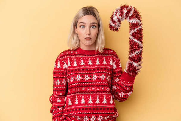 Young caucasian woman holding big Christmas stick isolated on yellow background shrugs shoulders and open eyes confused. - Photo, Image