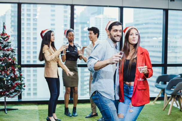 Pár kavkazských kancelářských pracovníků slaví Vánoce a Nový rok s cinknutím sklenice a pít víno spolu s šťastný a úsměv na pozadí mluvit o skupinových přátel ve společnosti. - Fotografie, Obrázek