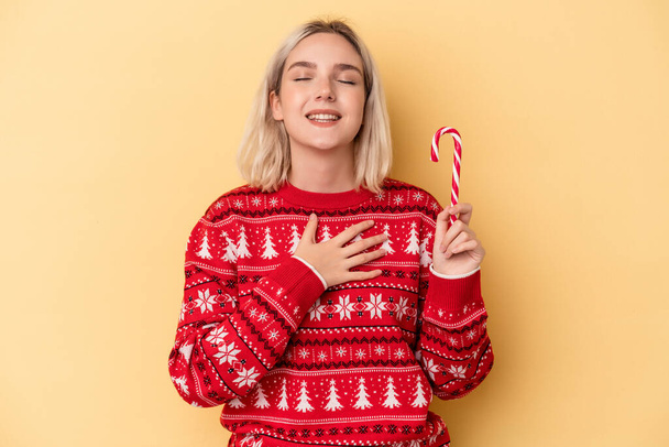 Young caucasian woman holding a christmas stick isolated on yellow background laughs out loudly keeping hand on chest. - Photo, Image