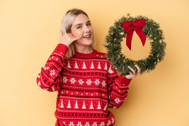 Young caucasian woman holding a Christmas wreath isolated on yellow background showing a mobile phone call gesture with fingers. - Foto, Imagem