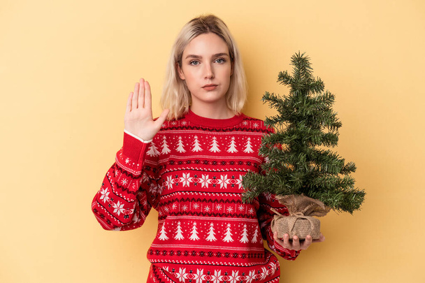 Jonge blanke vrouw met een kleine kerstboom geïsoleerd op gele achtergrond staan met uitgestrekte hand tonen stopteken, voorkomen dat u. - Foto, afbeelding