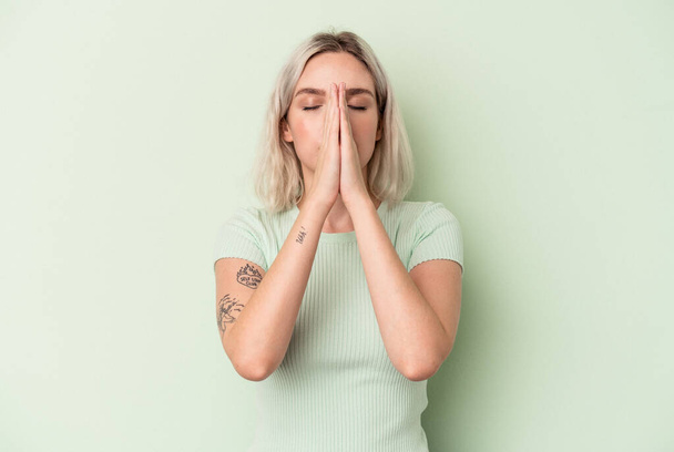 Young caucasian woman isolated on green background holding hands in pray near mouth, feels confident. - Photo, Image