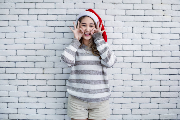 Portrait cute Asian young woman in costume Christmas and wear red santa claus hat with smile on white brick background and copy space. Happy of teen woman in theme Christmas. - 写真・画像