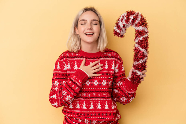 Young caucasian woman holding big Christmas stick isolated on yellow background laughs out loudly keeping hand on chest. - Photo, Image