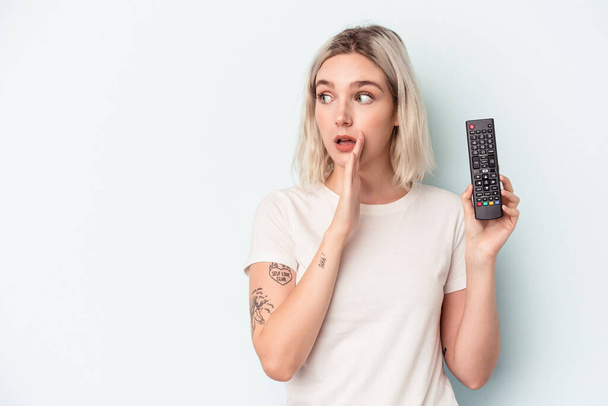 Young caucasian woman holding a tv controller isolated on blue background is saying a secret hot braking news and looking aside - Zdjęcie, obraz