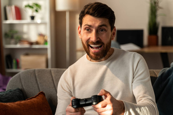 Excited young man playing video games and making winner gesture, while sitting at the sofa. Online competitions concept  - Photo, Image