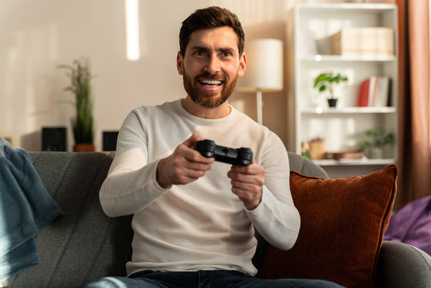 Waist up portrait of the bearded caucasian man with joystick playing video game alone on the couch. Stock photo  - Fotografie, Obrázek
