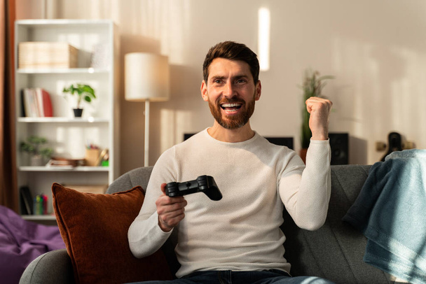 Yeah. Excited young handsome man playing video game and keeping mouth open while sitting on the couch at home  - Fotografie, Obrázek
