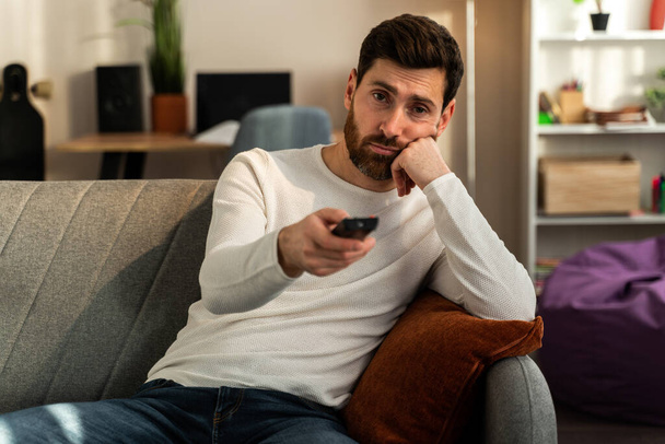 Photo of young bored man sitting on couch at home and changing TV channels with disinterest. Stock photo  - Fotó, kép