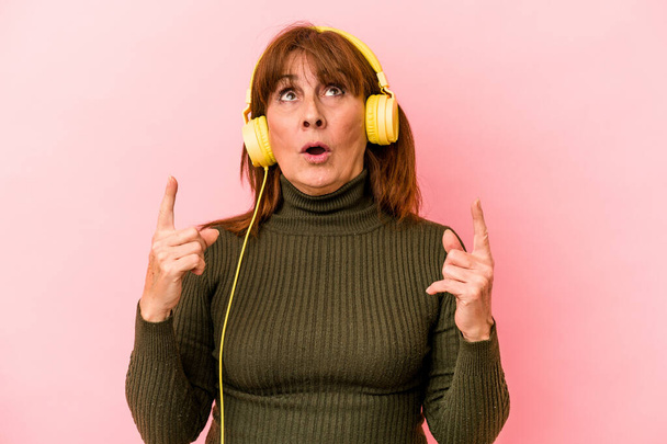 Middle age caucasian woman listening to music isolated on pink background pointing upside with opened mouth. - Foto, afbeelding