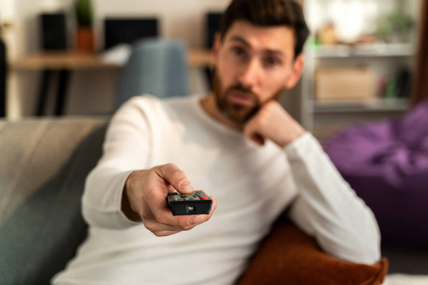 Aburrido joven caucásico hombre sosteniendo tv control remoto mientras está sentado en un sofá en casa mientras se relaja en el fin de semana  - Foto, imagen