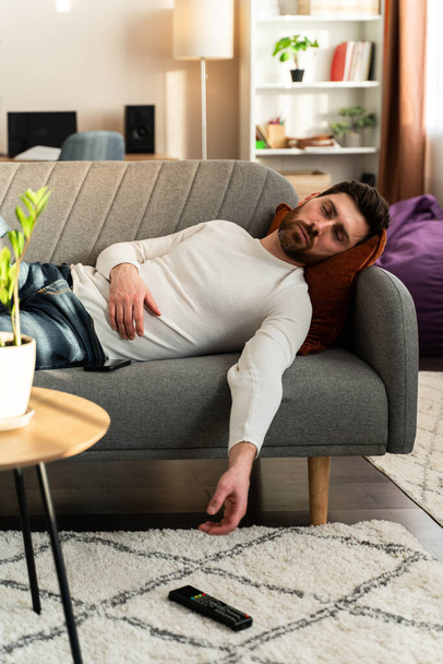 Bearded man sleeping on sofa with tv remote control at hands at home during the day. Leisure and people concept  - Photo, image
