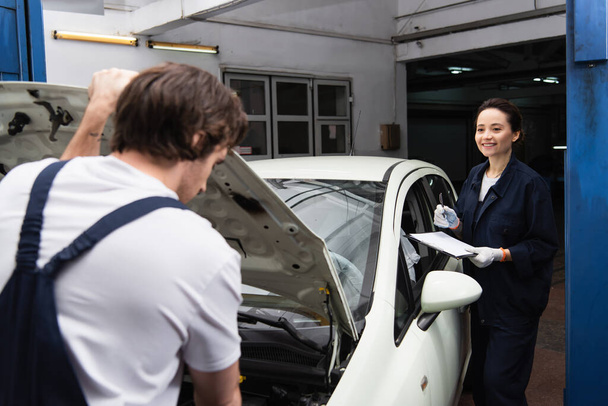 Trabajadora positiva sosteniendo portapapeles cerca borrosa colega y coche en servicio  - Foto, Imagen