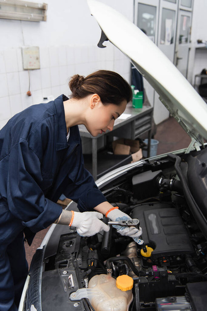 Side view of workwoman holding wrench near auto with open hood in garage  - Foto, afbeelding