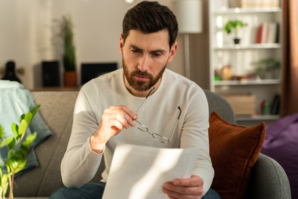 Frustrated millennial guy holding glasses and reading bad unpleasant news in postal letter. Sad confused young man considering paperwork and getting negative answer in post correspondence - Foto, Imagem