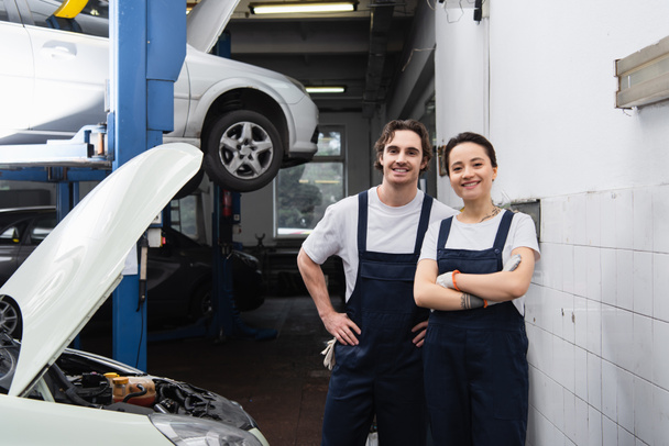 Cheerful mechanic in uniform looking at camera near car in garage - Photo, Image