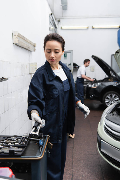 Young mechanic taking tool near auto and blurred colleague in garage  - Photo, Image