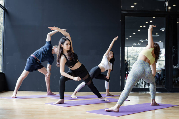 Fiatalok csoportja jógázik egy jóga matracon egy edzővel, aki fokozatosan tanít a tornateremben. Fiatal nők, férfiak és tanáraik jógáznak a tornateremben. A jóga általi testmozgás fogalma. - Fotó, kép