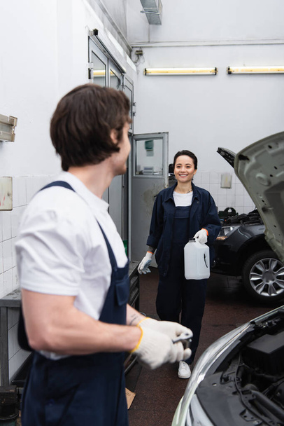 Usmívající se mechanik drží kanystr v blízkosti rozmazané kolegy a auto v provozu  - Fotografie, Obrázek