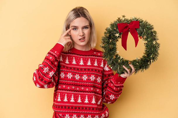Young caucasian woman holding a Christmas wreath isolated on yellow background showing a disappointment gesture with forefinger. - 写真・画像