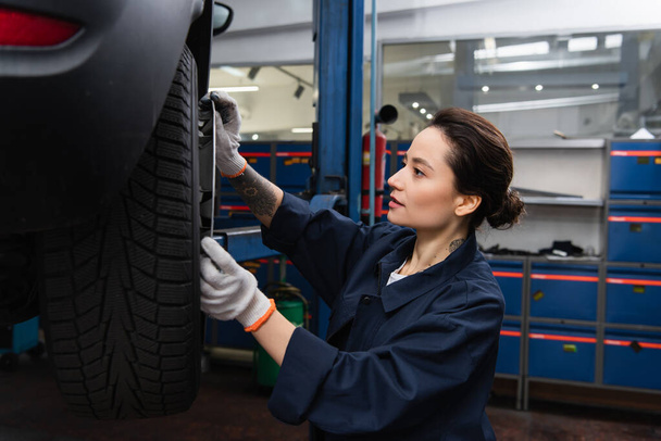 Young workwoman holding disk near wheel of blurred car in garage  - Zdjęcie, obraz