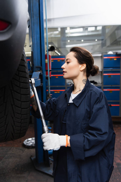 Side view of forewoman in uniform holding wrench near wheel of auto in garage  - Zdjęcie, obraz