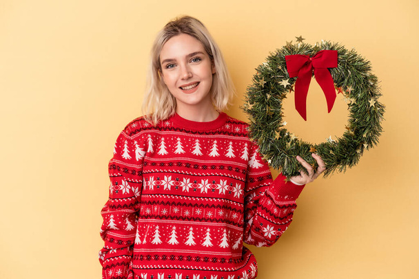 Young caucasian woman holding a Christmas wreath isolated on yellow background happy, smiling and cheerful. - 写真・画像